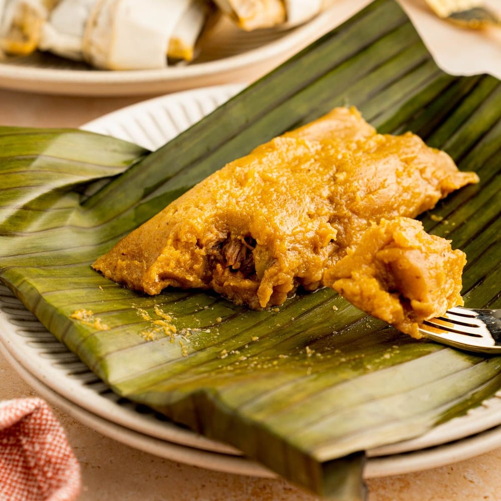 A pastel opened on a banana leaf, its pork filling showing, with more wrapped pasteles behind it.